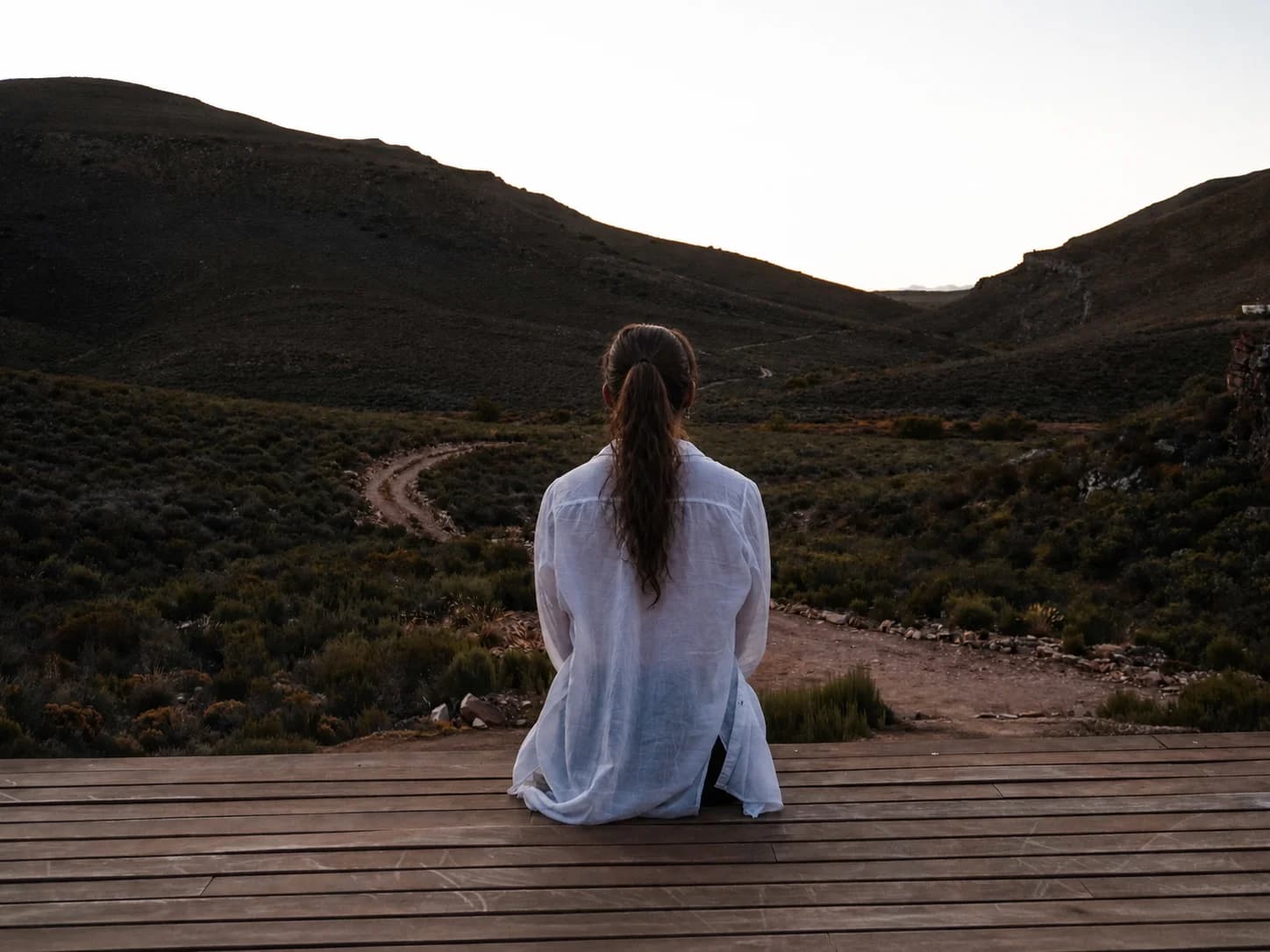Femme assise sur une terrasse en bois, contemplant un chemin qui serpente dans la vallée